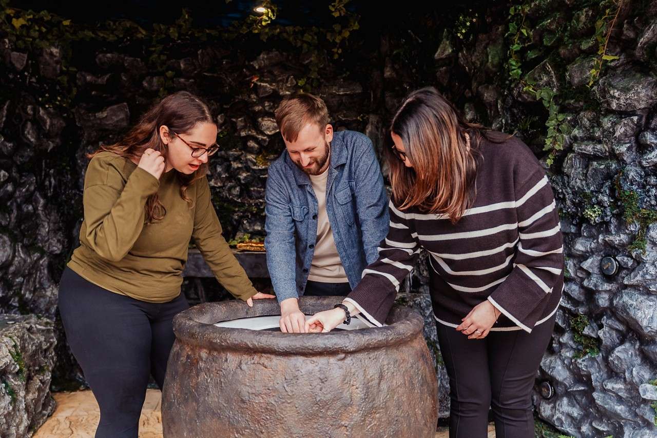 Three people examine papers inside a large stone basin, leaning in and pointing; they stand in a cave-like room with rocky walls and hanging ivy.