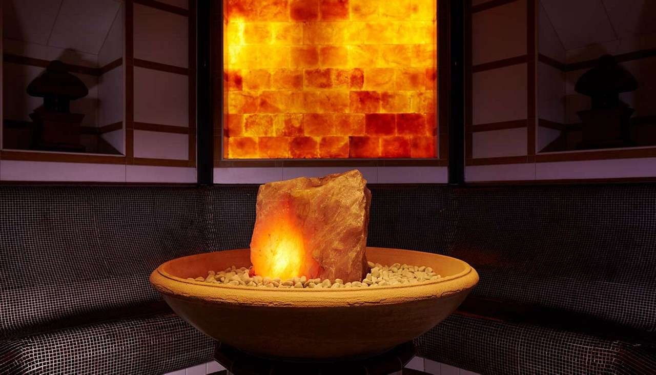 Glowing orange salt rock illuminates from a shallow round bowl filled with pebbles; surrounded by dark tiled curved bench; a backlit wall of orange salt bricks provides a warm backdrop.