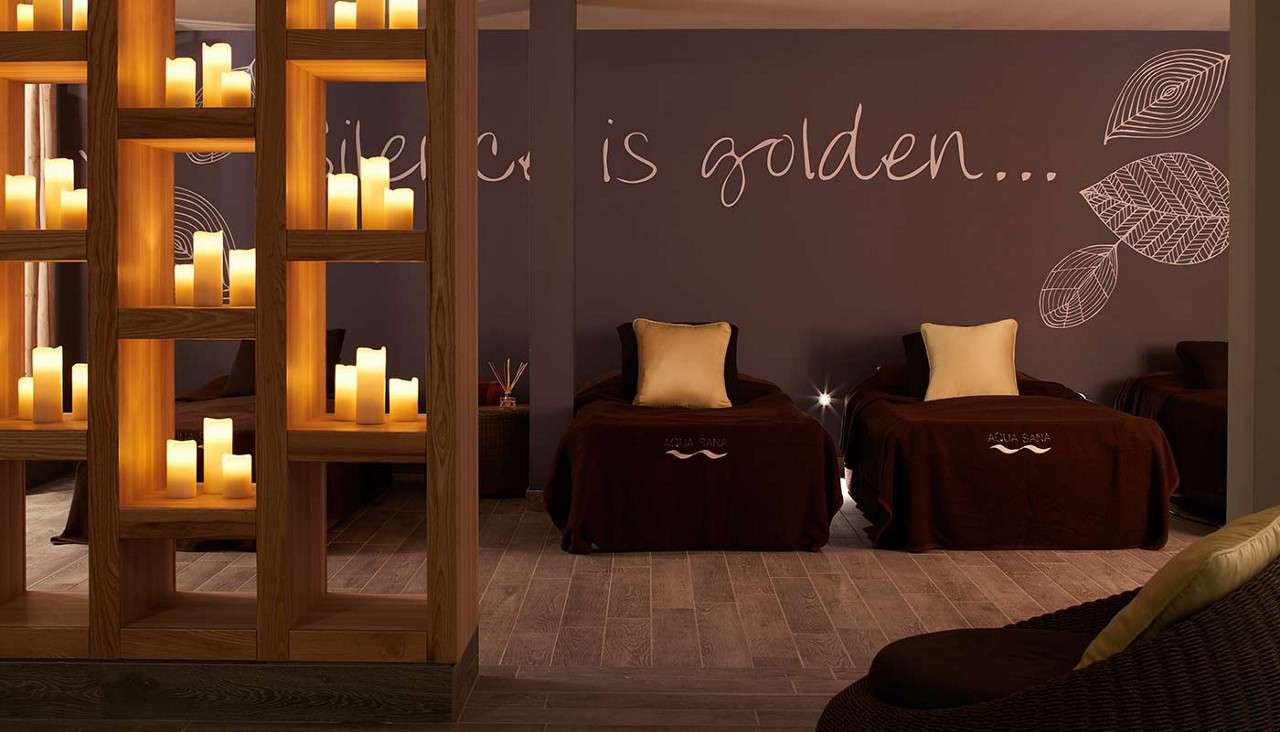 Candles glow on wooden shelves, softly lighting lounge beds with cream pillows in a spa relaxation room. Wall text: "silence is golden..." Blankets read "AQUA SANA" "AQUA SANA".