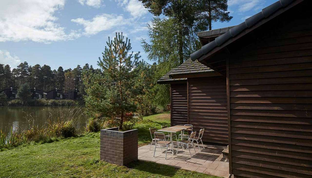 Patio table with four metal chairs sits on a small paved terrace, beside a wooden cabin, overlooking a calm lake bordered by trees under partly cloudy skies.