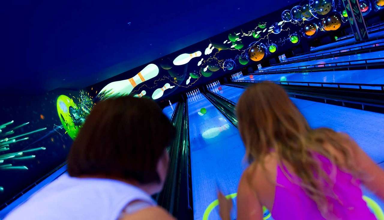 Two people bowl, releasing neon balls down glowing lanes in a blacklight bowling alley, with cosmic murals of pins and balls on the walls and multiple lanes stretching forward.