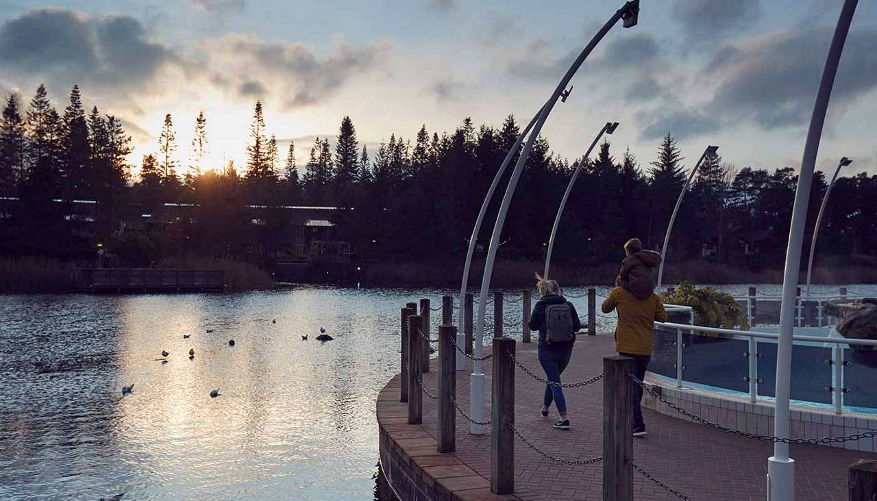 Two people on a curved lakeside promenade; one carries a child on their shoulders. Ducks float as sunset silhouettes pine trees. Arched light poles and chain posts frame the path.