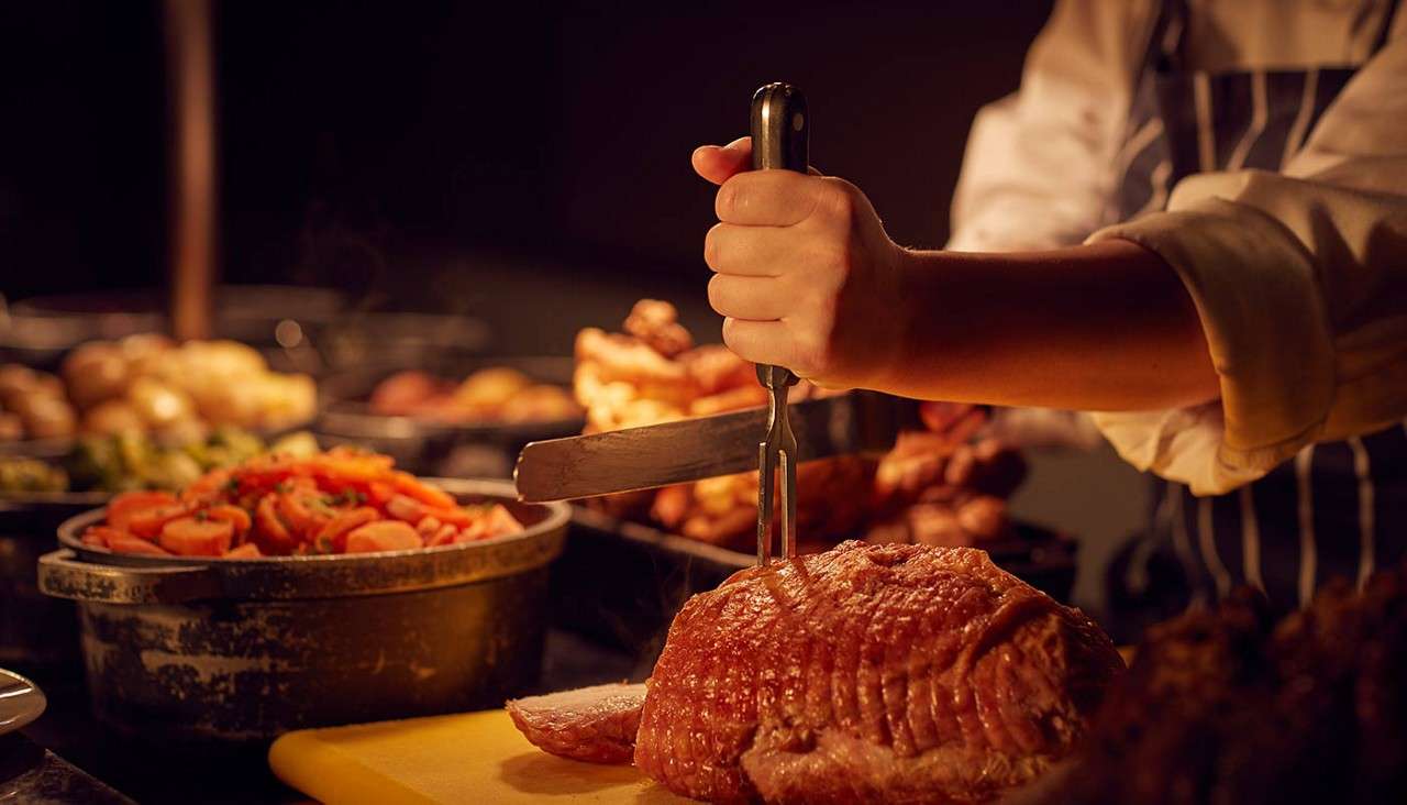Roast beef—held steady with a carving fork—gets sliced by a cook’s knife on a yellow board, surrounded by buffet dishes of carrots, potatoes, and greens under warm restaurant lighting.