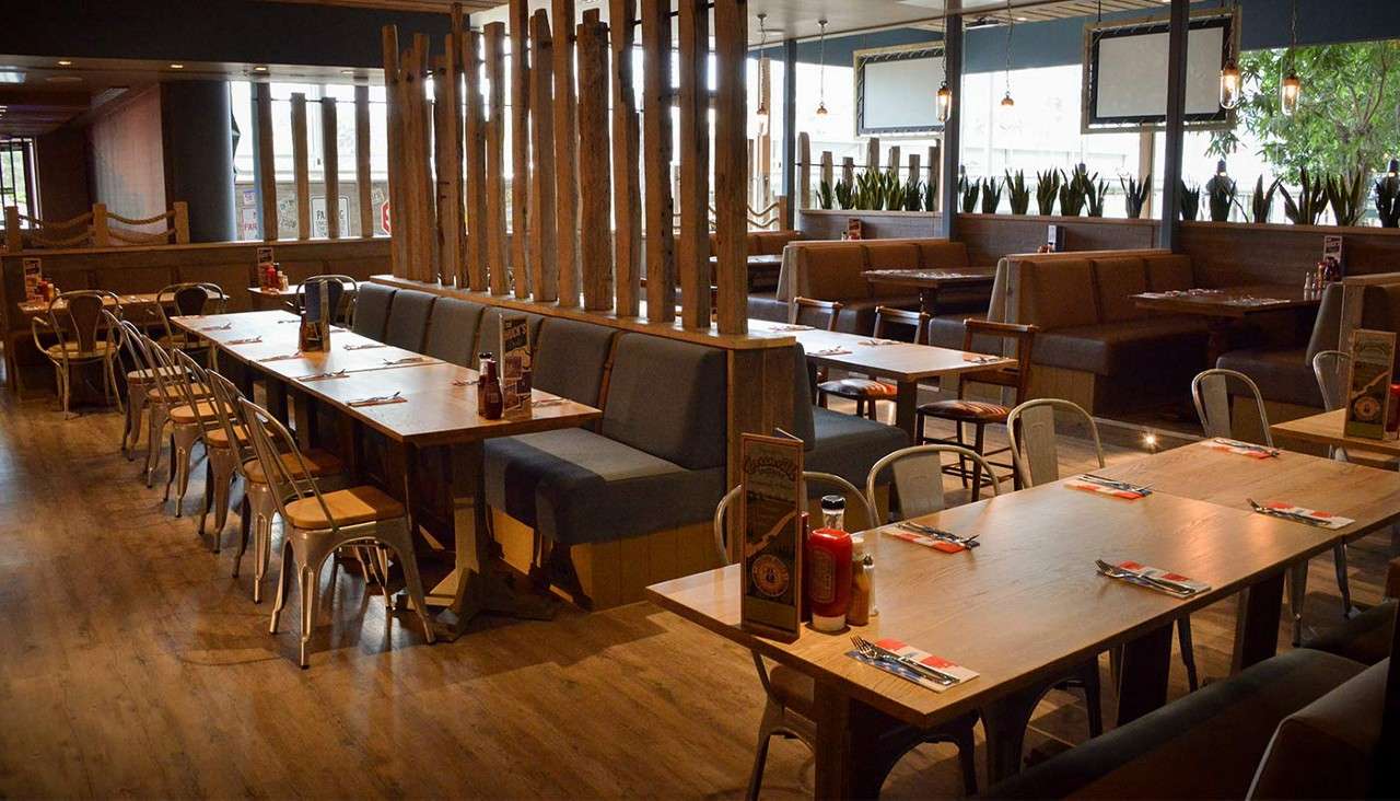 Dining tables set with cutlery and condiments line upholstered booths, while metal chairs surround them, in a spacious wood-floored restaurant featuring wooden partitions, potted plants, and pendant lights.