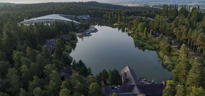 Lake reflects sky; small boats cluster at a dock. Surrounding context: dense pine forest with scattered cabins and a large glass-roof leisure complex, viewed from above in soft evening light.