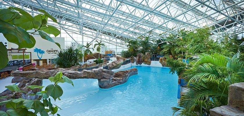 Indoor swimming pool rests beneath a glass atrium, bordered by tropical plants and faux-rock bridges; people absent. Context: waterpark with seating area and a sign reading: “CAFÉ CANOPY'S BAR”.