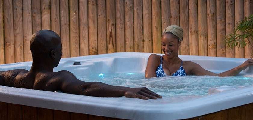 Two people relax and chat in a bubbling hot tub, leaning against the edges, surrounded by a wooden privacy fence and soft lights, in an outdoor evening setting.