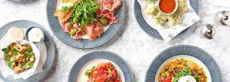 Assorted Mediterranean dishes sit arranged on blue-rimmed plates, featuring cured meats with greens, fried bites with dip, tomato salad, and chickpea salad, on a bright marble tabletop.