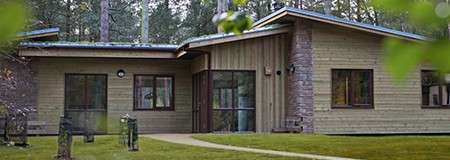House sits among pine trees; low, modern cabin with wood siding and stone accents. Windows reflect forest; a curved path leads across trimmed lawn; soft light suggests overcast woodland setting.
