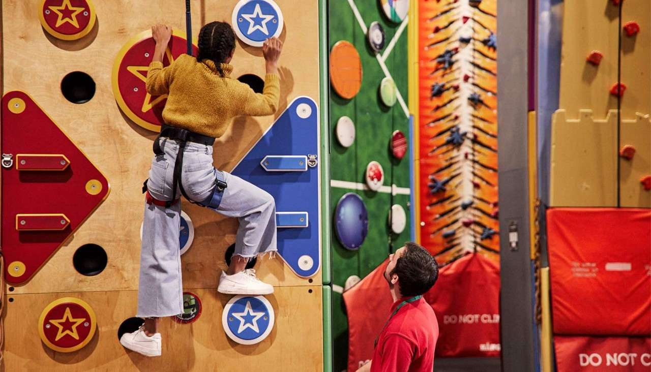 Climber wearing a harness ascends a colorful indoor wall, gripping star-marked holds; instructor watches below beside padded mats. Text visible: DO NOT CL, DO NOT CL.