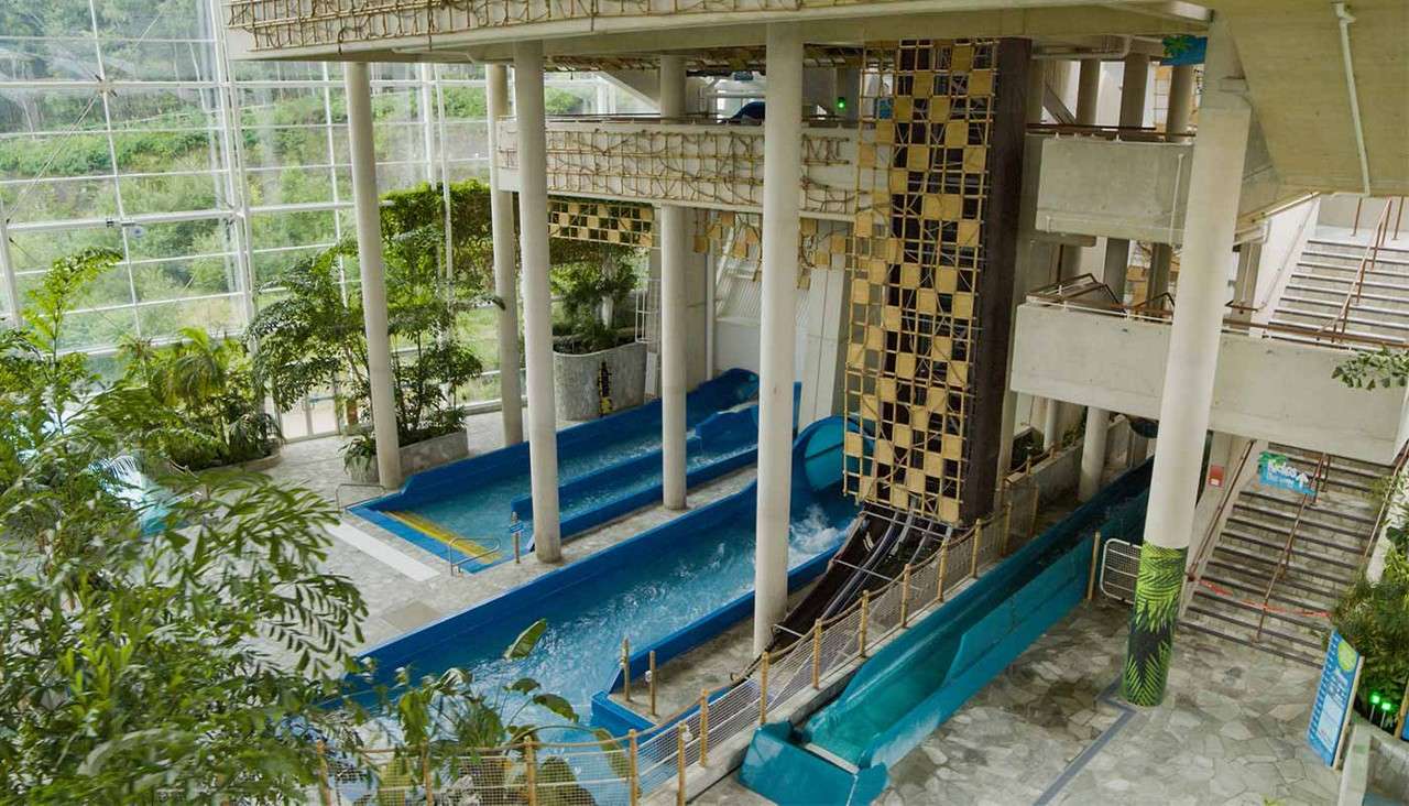 Blue water slide channels discharge foaming water into shallow run-outs, beneath concrete columns and rope bridges; surrounding context includes indoor tropical plants, glass walls, and stairways in a spacious atrium.