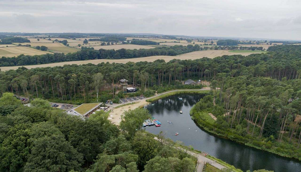 Small lake hosts pedal boats and kayaks; people gather on a sandy beach and paths. Pine forest and buildings encircle the water, with farmland stretching beyond under overcast skies.