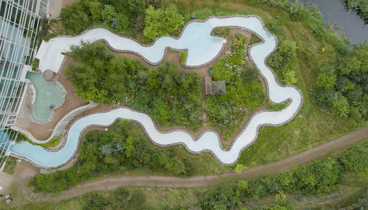 Winding artificial water channel curves through a landscaped garden. It loops around dense greenery and a small gazebo, beside a glass-roofed building, dirt paths, and a nearby natural waterway.