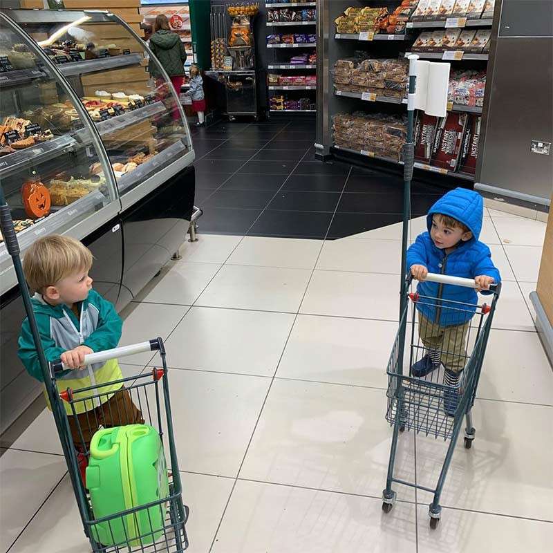 Two small children push miniature shopping carts, facing each other, pausing near a bakery display and bread shelves in a supermarket, one in a blue hooded jacket, the other green.