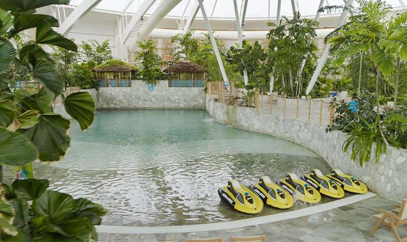 Yellow personal watercraft labeled “SEA-DOO” sit idle at the edge of a calm pool, surrounded by lush tropical plants, stone walls, and thatched cabanas beneath a bright, vaulted indoor roof.