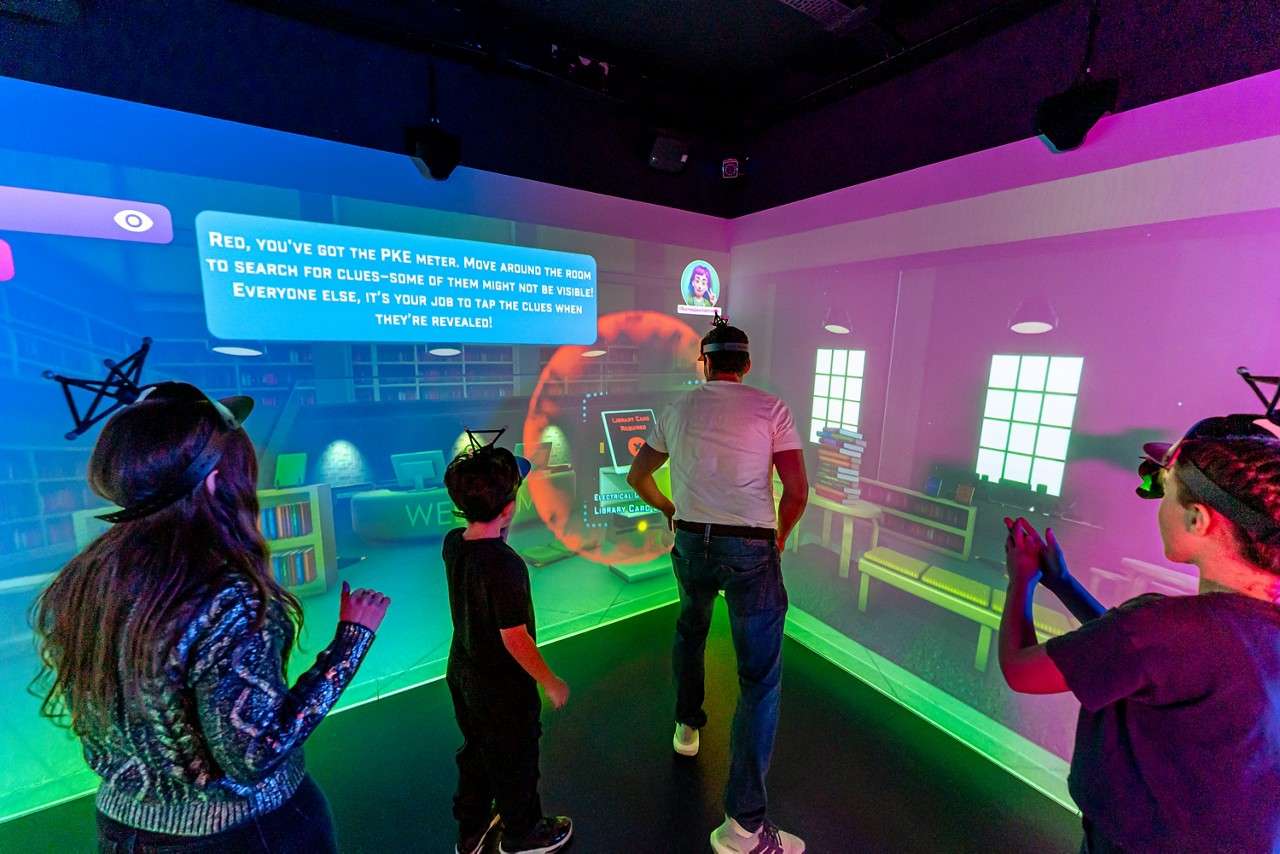 Players wearing headsets interact with a projected game wall in a neon-lit room. On-screen text: "RED, YOU'VE GOT THE PKE METER. MOVE AROUND THE ROOM TO SEARCH FOR CLUES—SOME OF THEM MIGHT NOT BE VISIBLE! EVERYONE ELSE, IT'S YOUR JOB TO TAP THE CLUES WHEN THEY'RE REVEALED!"