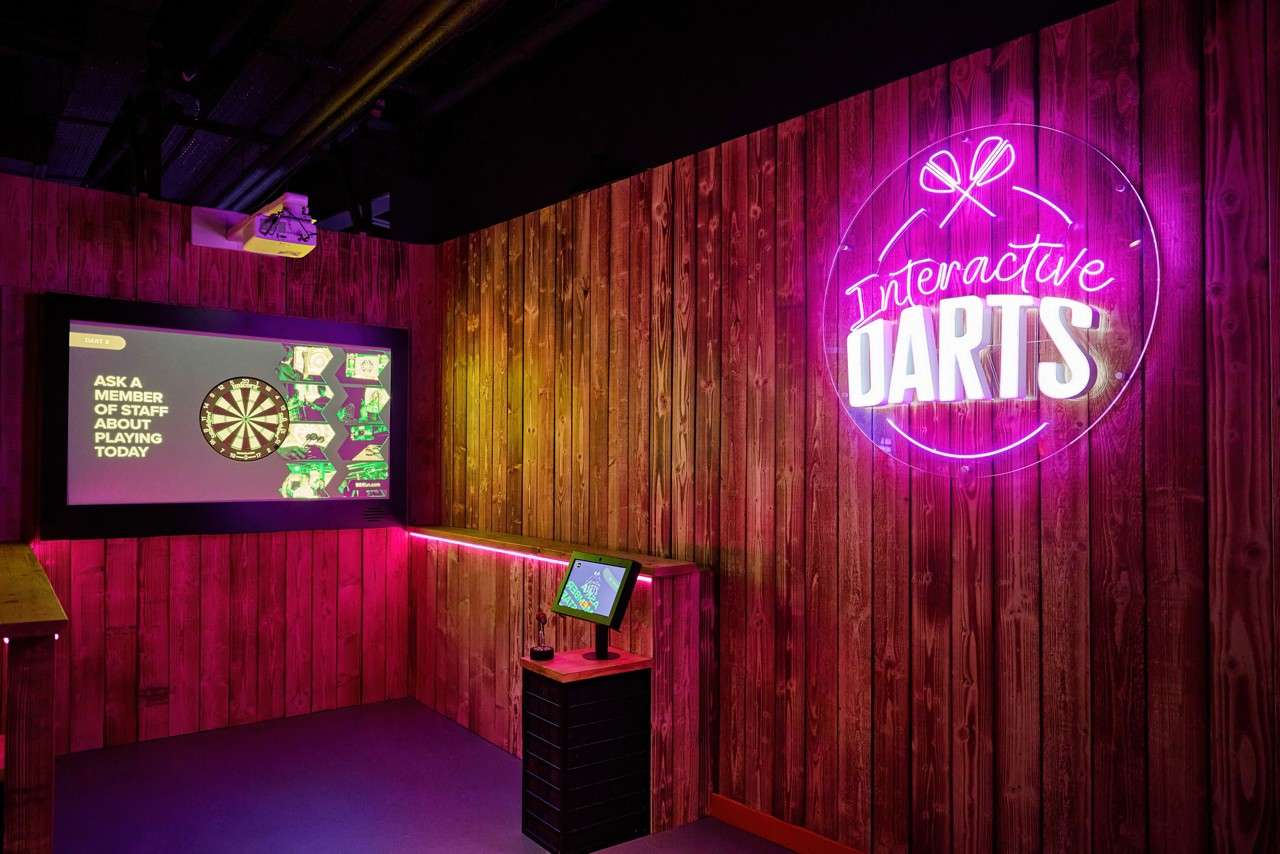 Digital dartboard screen displays instructions in a wood‑paneled gaming bay, while a neon sign glows and a small touchscreen kiosk stands nearby. Text: Interactive DARTS; ASK A MEMBER OF STAFF ABOUT PLAYING TODAY.