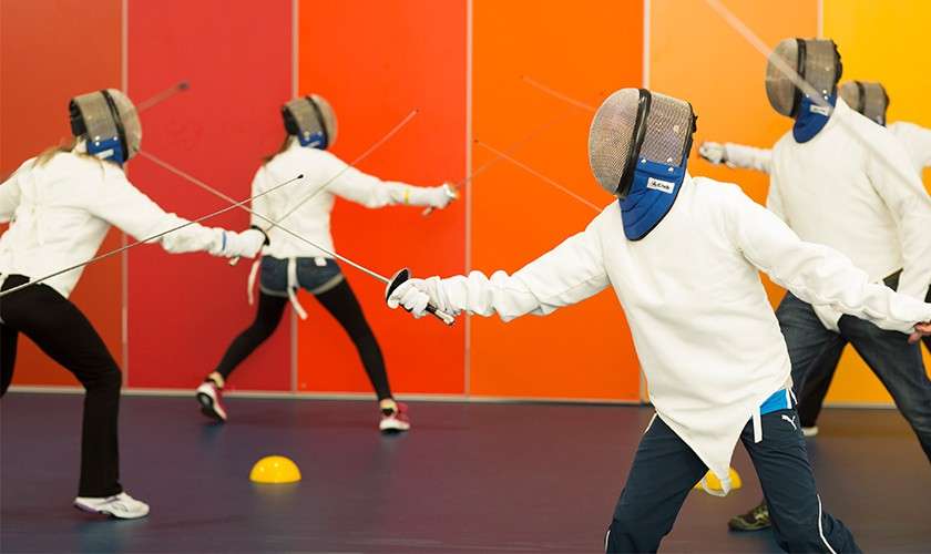 Fencers lunge, extending foils, wearing masks and white jackets; a group class practices footwork in a gym with blue floor, orange gradient wall, and small training cones.