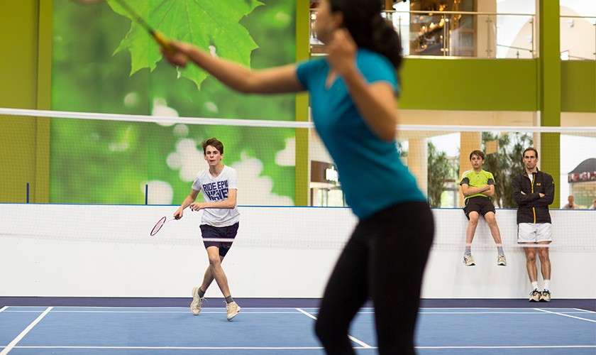 A mother and son are playing badminton on our indoor badminton court with the father and son watching at the side.