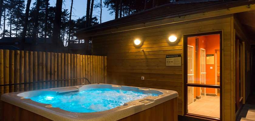 Hot tub bubbles and steams, illuminated blue, beside a wooden cabin. Outdoor wall lights glow, an open doorway reveals a lit interior, while a fence and twilight forest surround.