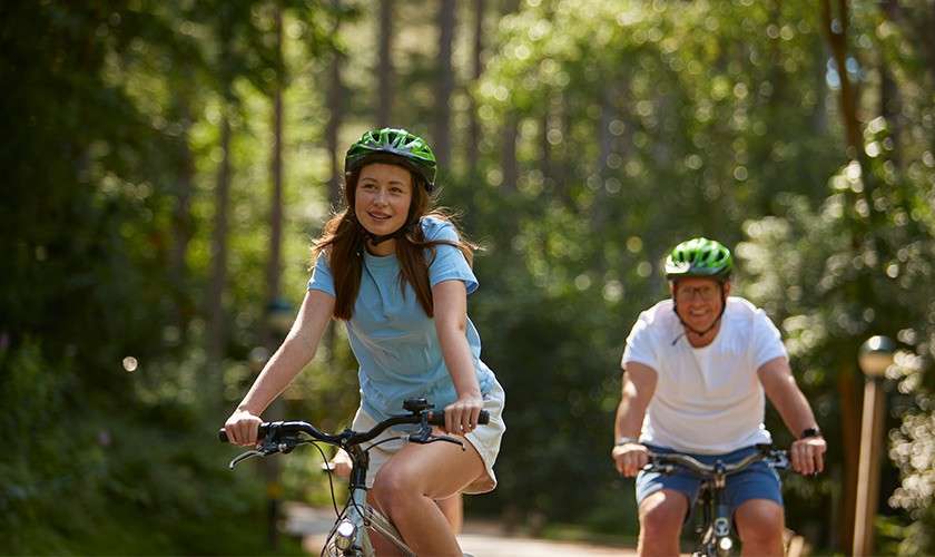 Two cyclists pedal and smile, wearing green helmets. They ride along a sunlit, tree-lined forest path, with one leading and another following slightly behind.