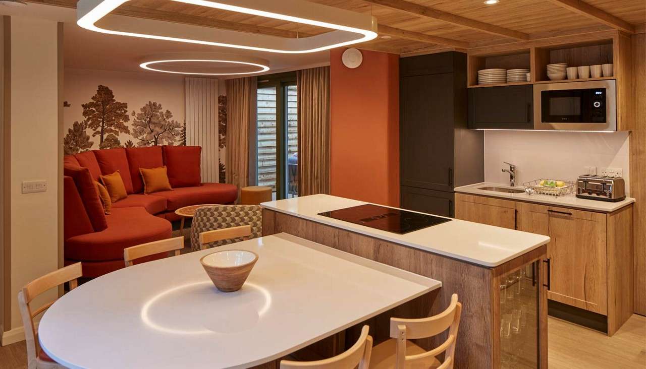 Kitchen island anchors cooking, with induction hob nearby, while halo lights glow overhead; context: a warm open-plan room with wooden cabinetry, dining table, and curved red sectional beside tree mural.
