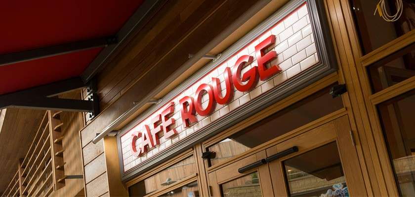 Signboard displays “CAFE ROUGE” in bold red letters, mounted above wooden-framed glass doors, viewed at an angle beneath a canopy, with a white tiled backdrop on the café exterior.