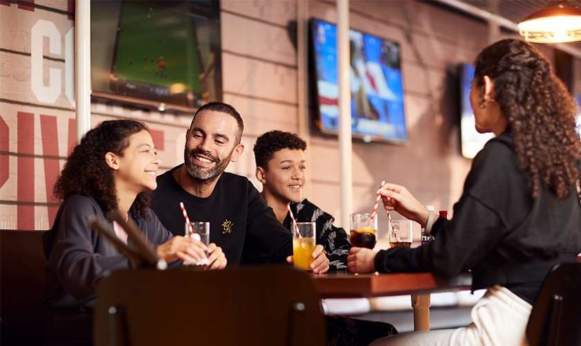 Four people sit at a restaurant table, smiling and chatting while drinking sodas with striped straws. Televisions play sports on the wall behind them in a warmly lit casual setting.