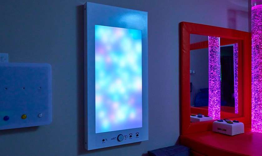 Illuminated sensory wall panel glows with soft blue and pink light patterns, while adjacent bubble tubes shine magenta; padded mirror, bench, and small control box create a calming therapy room.