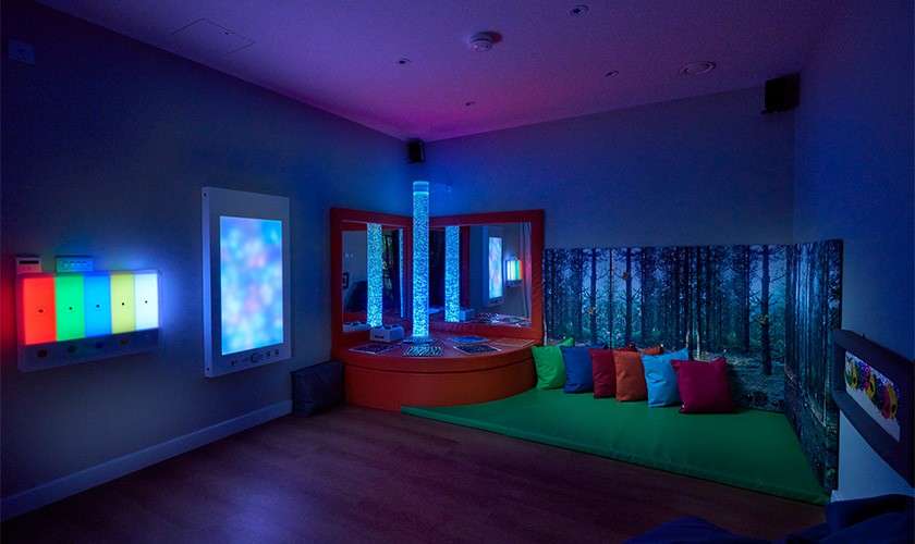 Multisensory room—glowing bubble tubes and LED panels illuminate. In a dim space: mirrors, a padded platform, green mat with colorful cushions, forest-wall mural, and wooden floor create a calming therapeutic environment.