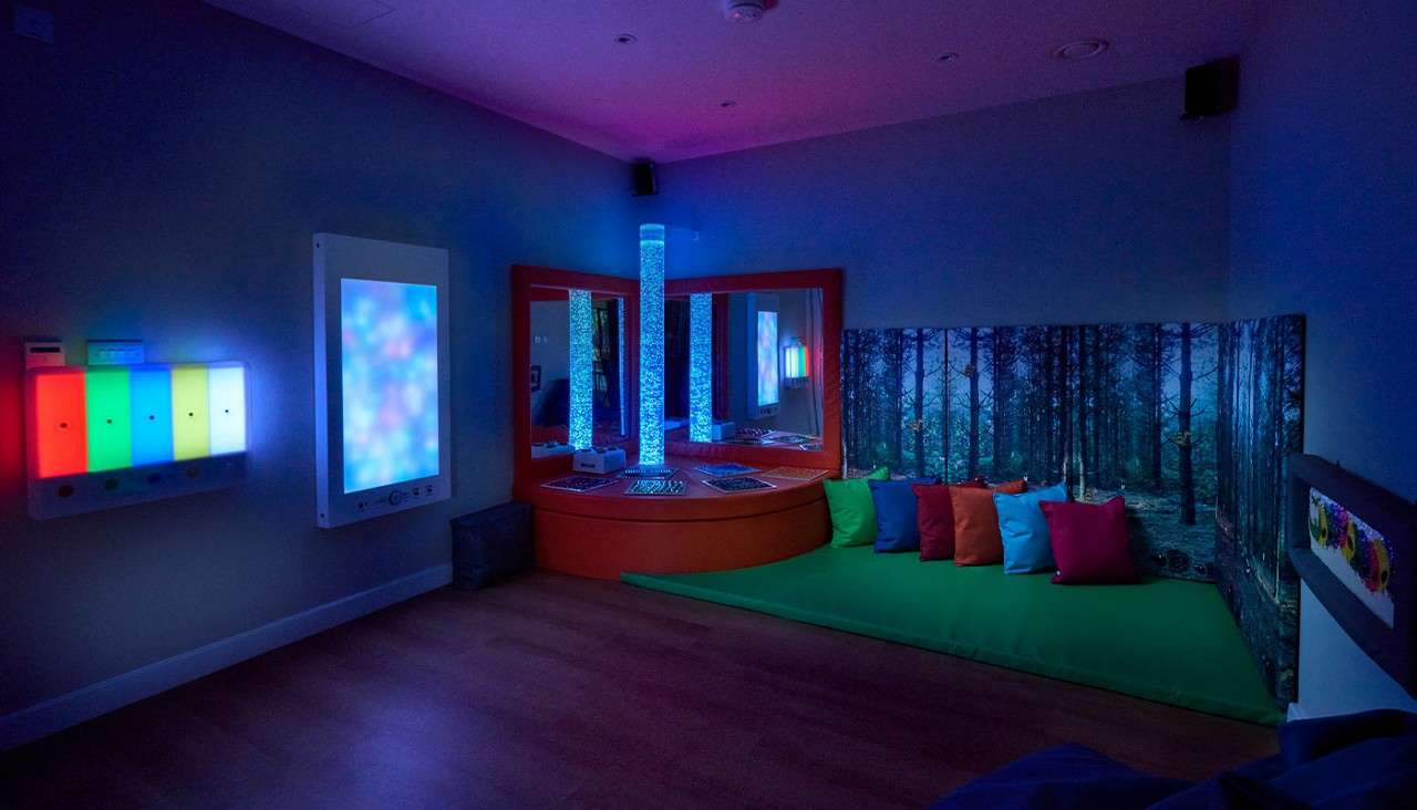 Two illuminated bubble tubes glow blue, reflecting in mirrors, while LED panels pulse multicolors. Context: a dim sensory room with a green mat, colorful cushions, and a forest-wall mural.