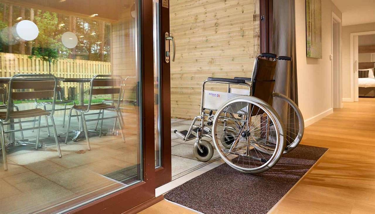 Wheelchair rests beside an open sliding glass door, bridging interior wood floor and outdoor patio; metal chairs and a wooden fence outside. Visible text on the wheelchair: ROMA.