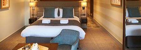 A neatly made double bed with folded towels sits between two lamps. Pillows accent it. An upholstered chair and tea tray sit foreground; hallway and wardrobe frame the hotel room.