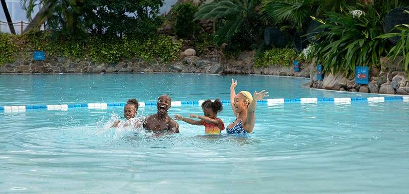 Family of four splashes and laughs, waist-deep, tossing water. Shallow outdoor pool encircled by a blue-and-white lane rope, stone edging, dense tropical greenery, and distant rocks under cloudy light.