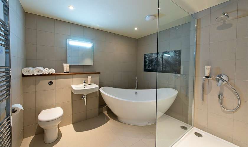 Freestanding white bathtub sits centered, surrounded by a modern bathroom: glass-walled shower, wall-mounted sink and mirror, toilet, heated towel rail, recessed lights, beige tiled walls, wooden shelf with towels.