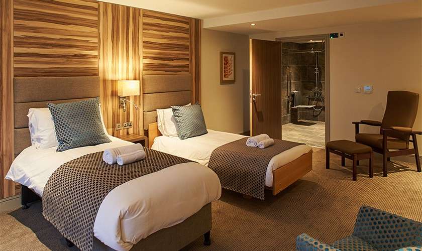 Twin beds stand side by side, topped with pillows and towels. Lighting and wooden headboards frame the room; a door shows an accessible bathroom with grab bars and shower seat.