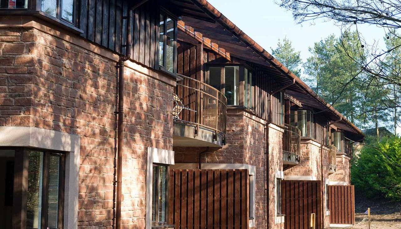 Row of stone-and-wood townhouses displays curved metal balconies and bay windows, catching dappled sunlight, in a forested residential setting with trees and shadows lining the exterior walkway.