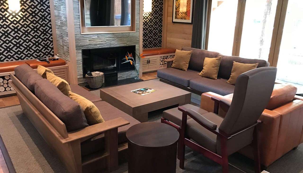 Sectional sofas and an armchair encircle a low square coffee table, facing a stone fireplace and wall-mounted TV; benches, patterned walls, log storage, and glass doors frame the living room.