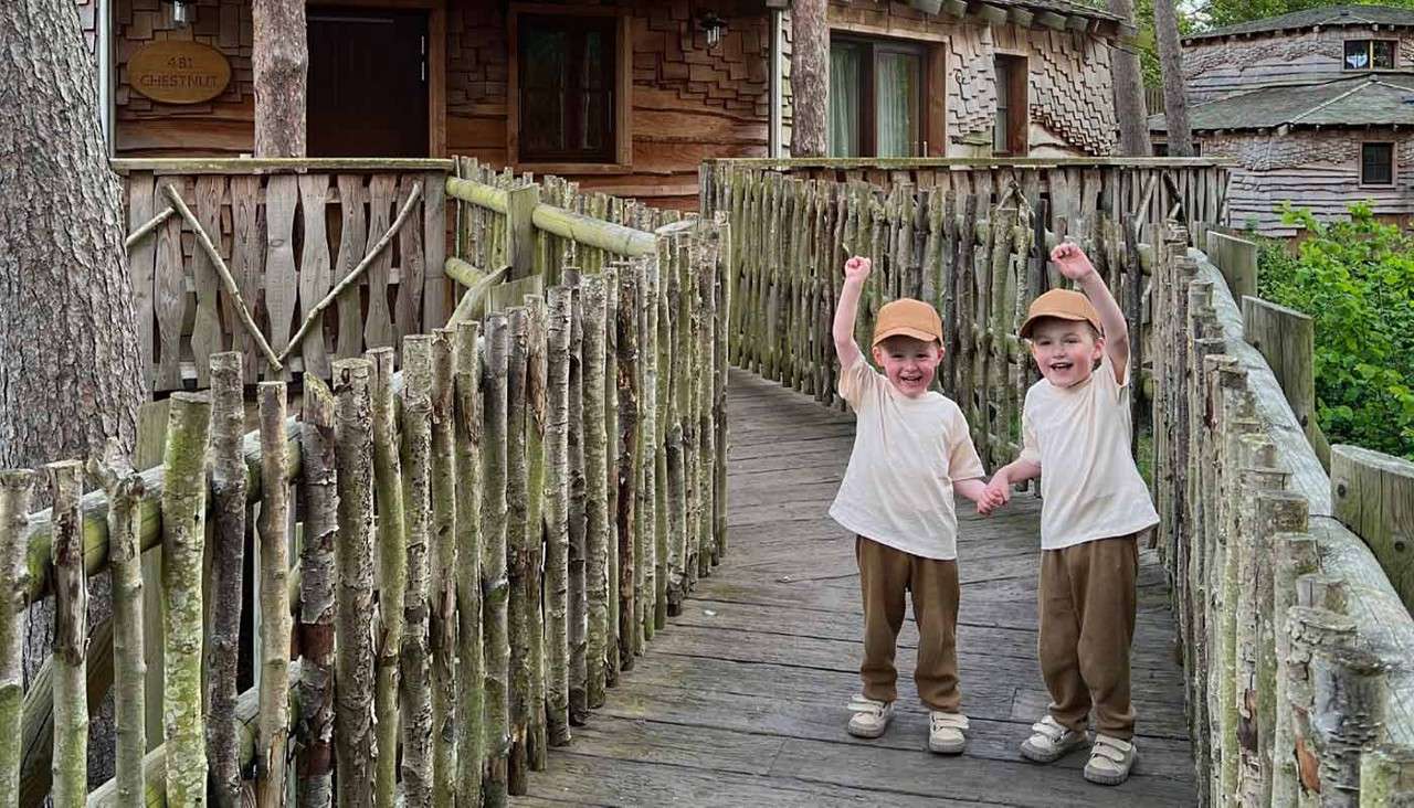Two young children hold hands and cheer, raising arms while standing on a rustic wooden footbridge. Surrounding treehouse cabins and railings made of branches create a forest setting. Text: 81 CHESTNUT.