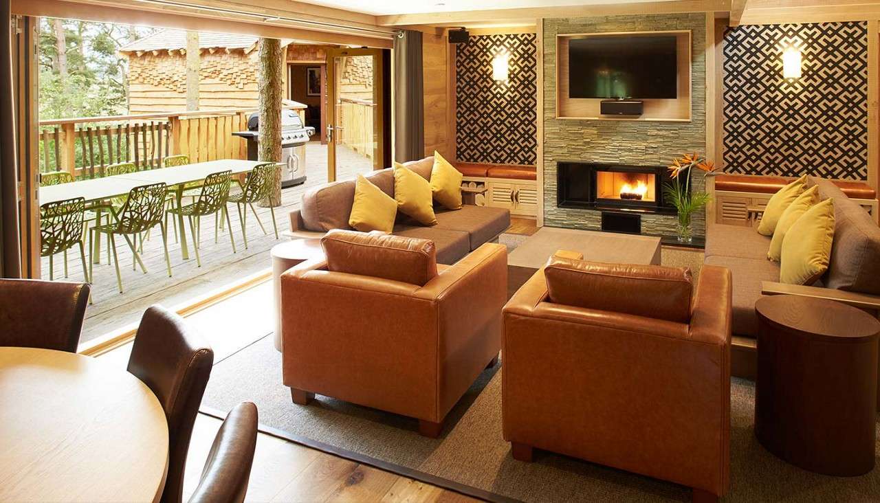 Living room opens onto a deck, revealing green chairs and a grill outside; inside, leather armchairs face a fireplace and TV, with sofas, coffee table, patterned walls, and forest setting.