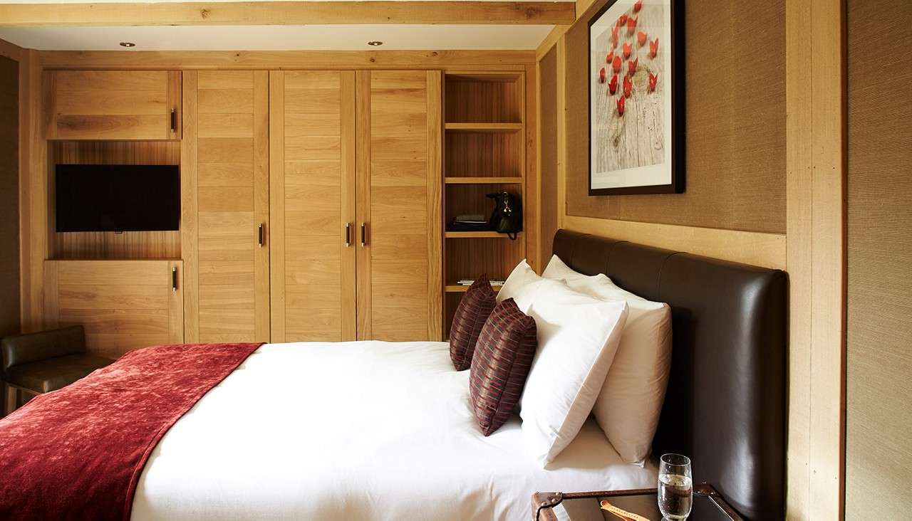 Bed lies neatly made, topped with red throw and striped pillows, against leather headboard; nearby are wooden built-in wardrobe, open shelves, TV, framed art, and bedside table with water glass.