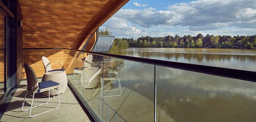 Balcony chairs face outward as a glass-railed terrace curves along a wooden-clad building, overlooking a calm lake with a distant tree line under a partly cloudy sky.