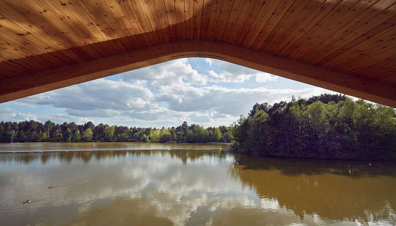 Wooden roof canopy frames a calm lake, sheltering the viewpoint; gentle ripples reflect clouds while a few ducks glide across; forested shoreline and partly cloudy sky form the serene background.