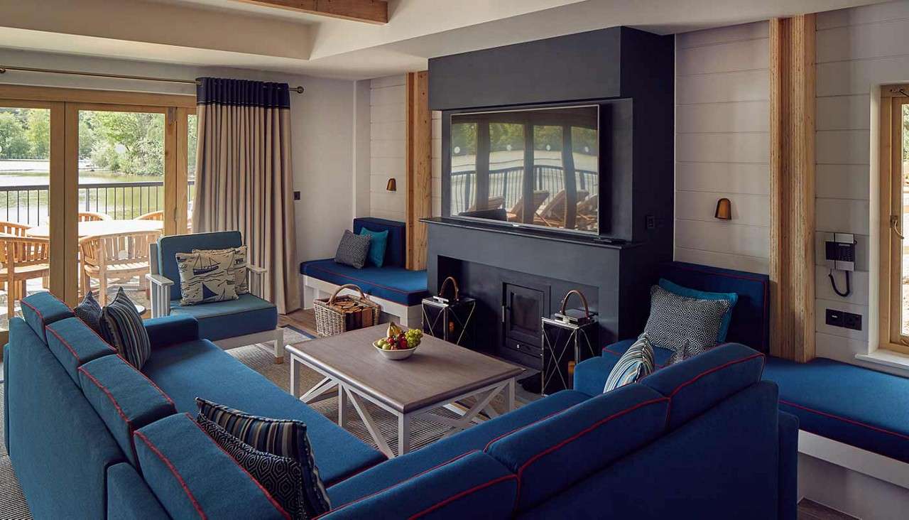 Blue sectional sofas surround a coffee table facing a large TV above a black fireplace, in a lakeview living room with sliding doors, balcony seating, built-in benches, cushions, and lanterns.