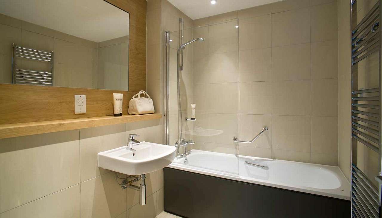 Bathtub anchors, fitted with glass screen and chrome showerhead; adjacent sink and wide mirror reflect light; beige tiled walls, wooden shelf, and heated towel rail complete the compact bathroom.