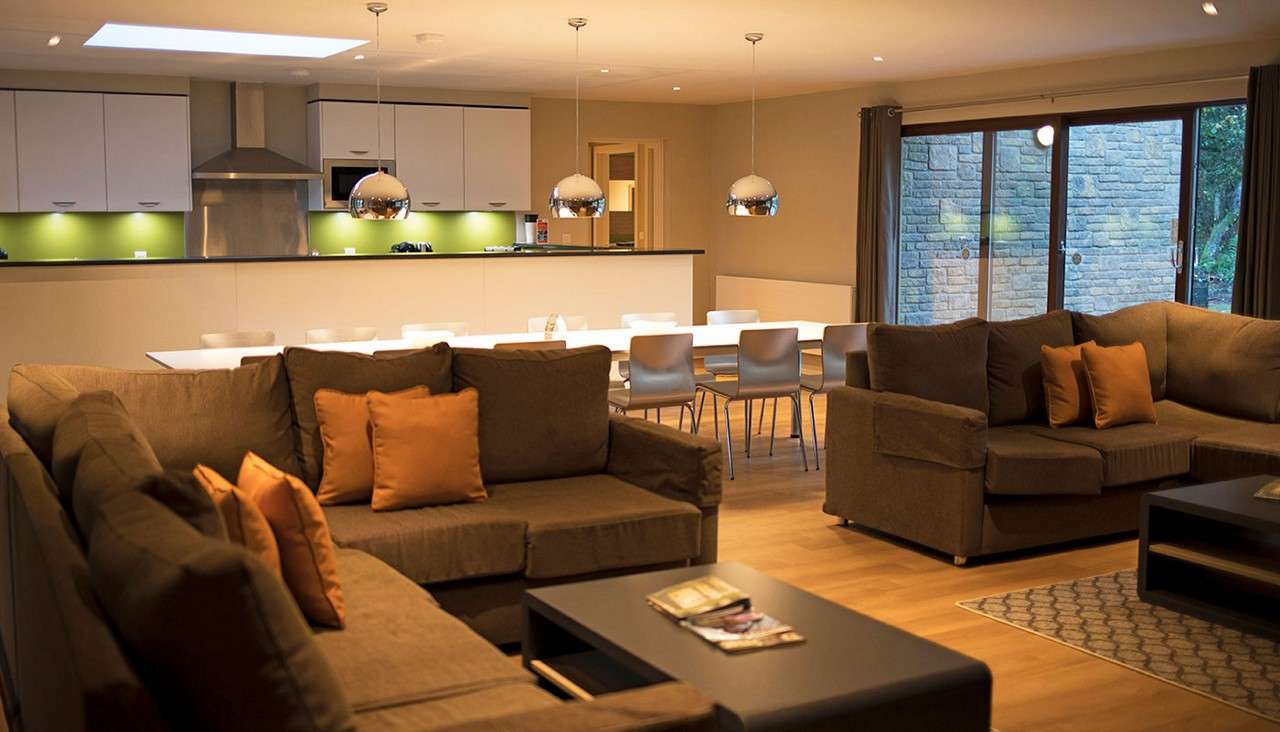 Sectional sofas frame a coffee table; pendant lights glow overhead. Context: an open-plan kitchen with green backsplash and long dining table, beside sliding glass doors and wood floors.