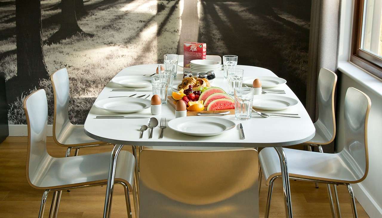 Dining table set with plates, glasses, cutlery, and egg cups waits for diners; fruit platter and pastries centered; six chairs surround it in a sunlit room with forest-mural wall.