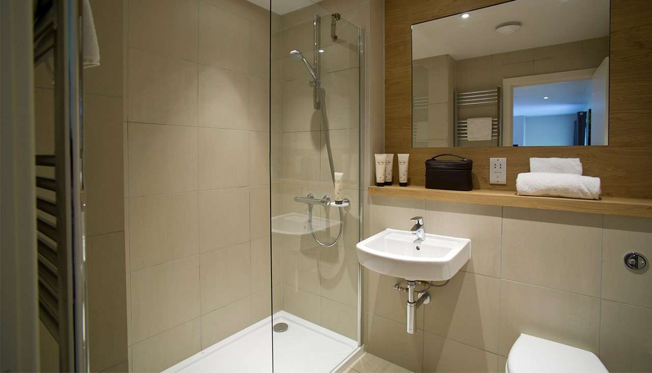 Glass-walled shower stands against beige tiles. Nearby, a wall-mounted sink sits beneath a large mirror and shelf holding toiletries and towels, in a modern bathroom with a toilet.