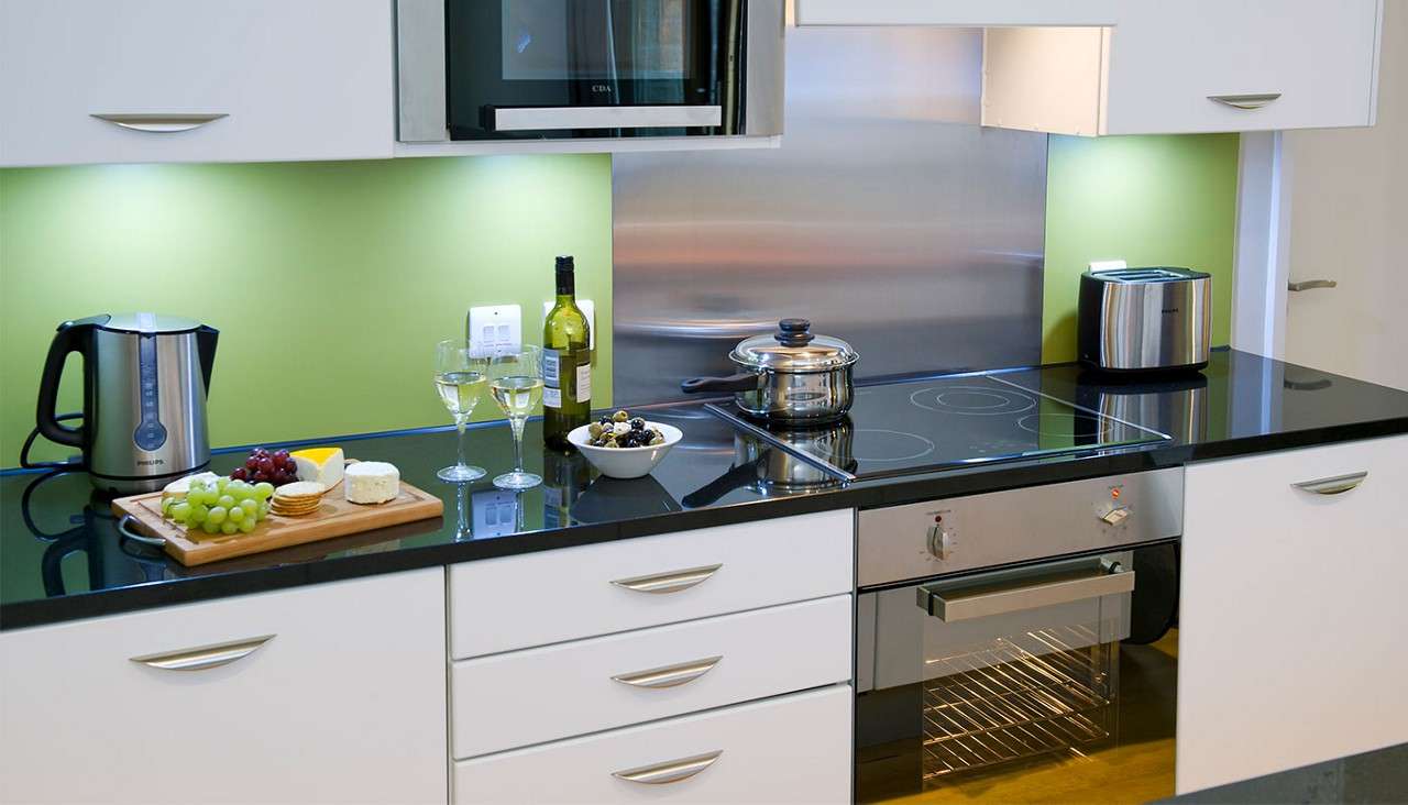 Countertop presents snacks and cookware in a modern kitchen; grapes, cheese, crackers, wine bottle and two glasses, bowl of olives, lidded pot on induction hob, kettle, toaster, microwave, oven. Visible text: PHILIPS, PHILIPS, CDA.