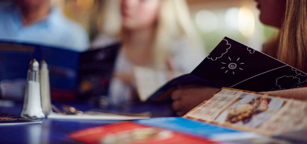 People hold and read menus; salt and pepper shakers and colorful menus rest on a table in a softly lit restaurant, with background diners blurred.